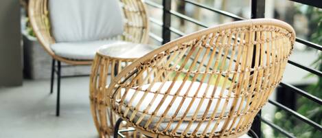 Two wicker chairs with white cushions on a balcony with a glass railing, overlooking a garden.