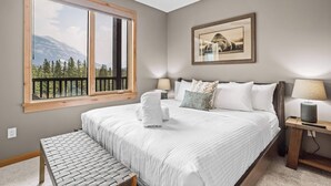 A bedroom with a large bed, a bench, a nightstand, a lamp, and a framed picture on the wall. - Spring Creek Vacations 1 Bedroom Elevated Condo 405TM (Canmore)
