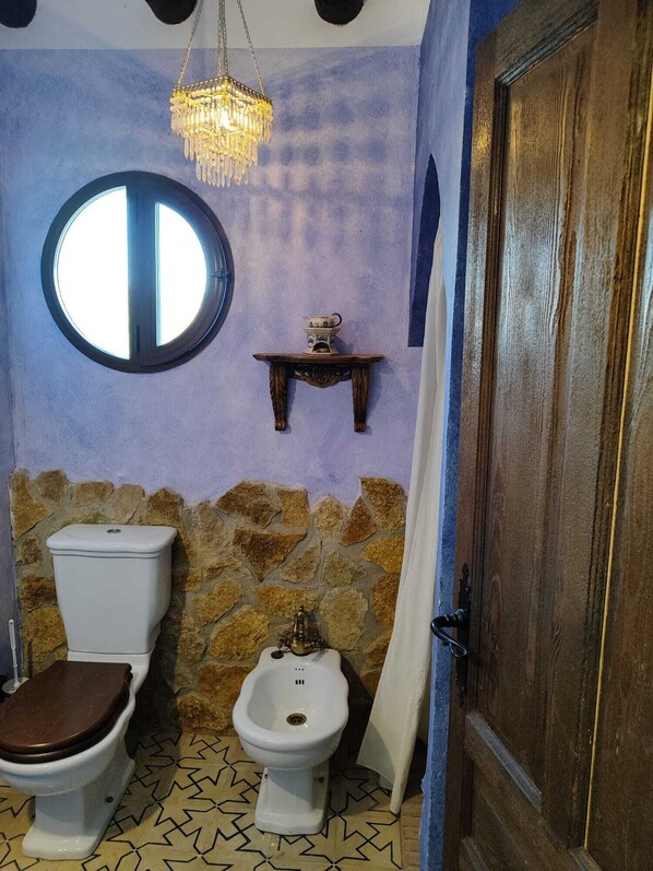 A bathroom with a stone wall, a wooden door, a round mirror, a toilet, a bidet, and a hanging light fixture. - Villa El Membrillar (Zagrilla Baja)