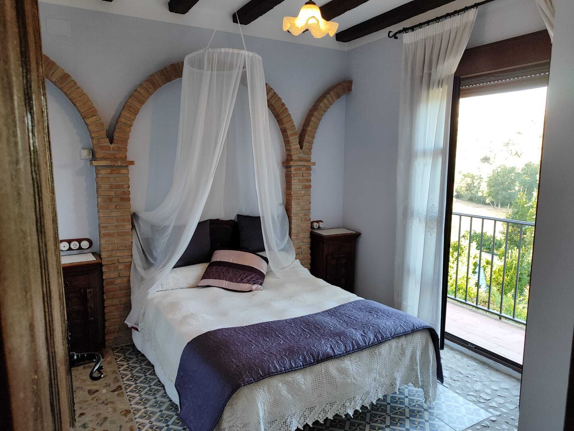 A bedroom with a bed under a canopy, brick arches, a balcony with a view, and a patterned floor.