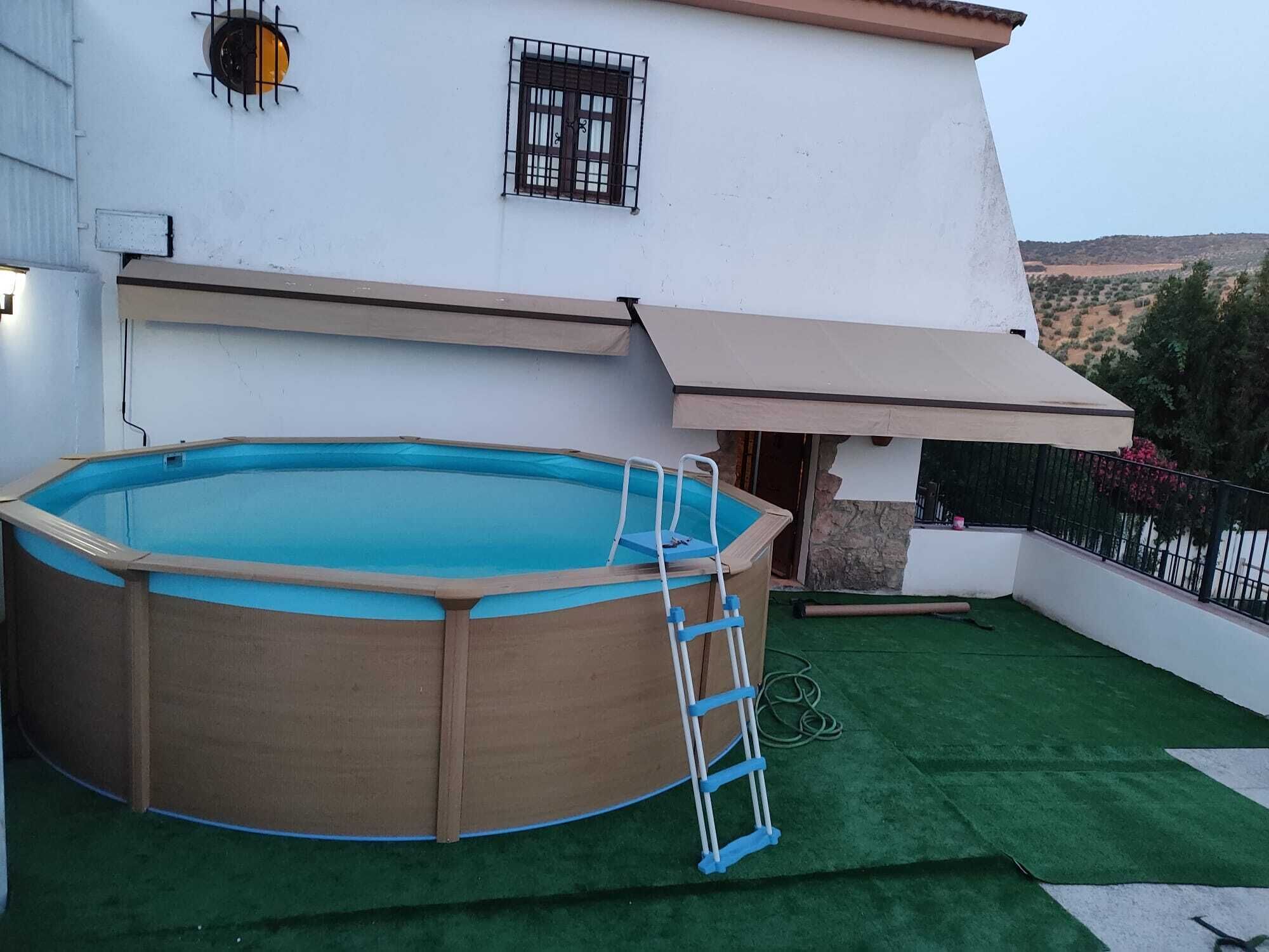 Une piscine extérieure avec un escalier, un bâtiment blanc doté d’une marquise marron et une zone recouverte de gazon synthétique vert.