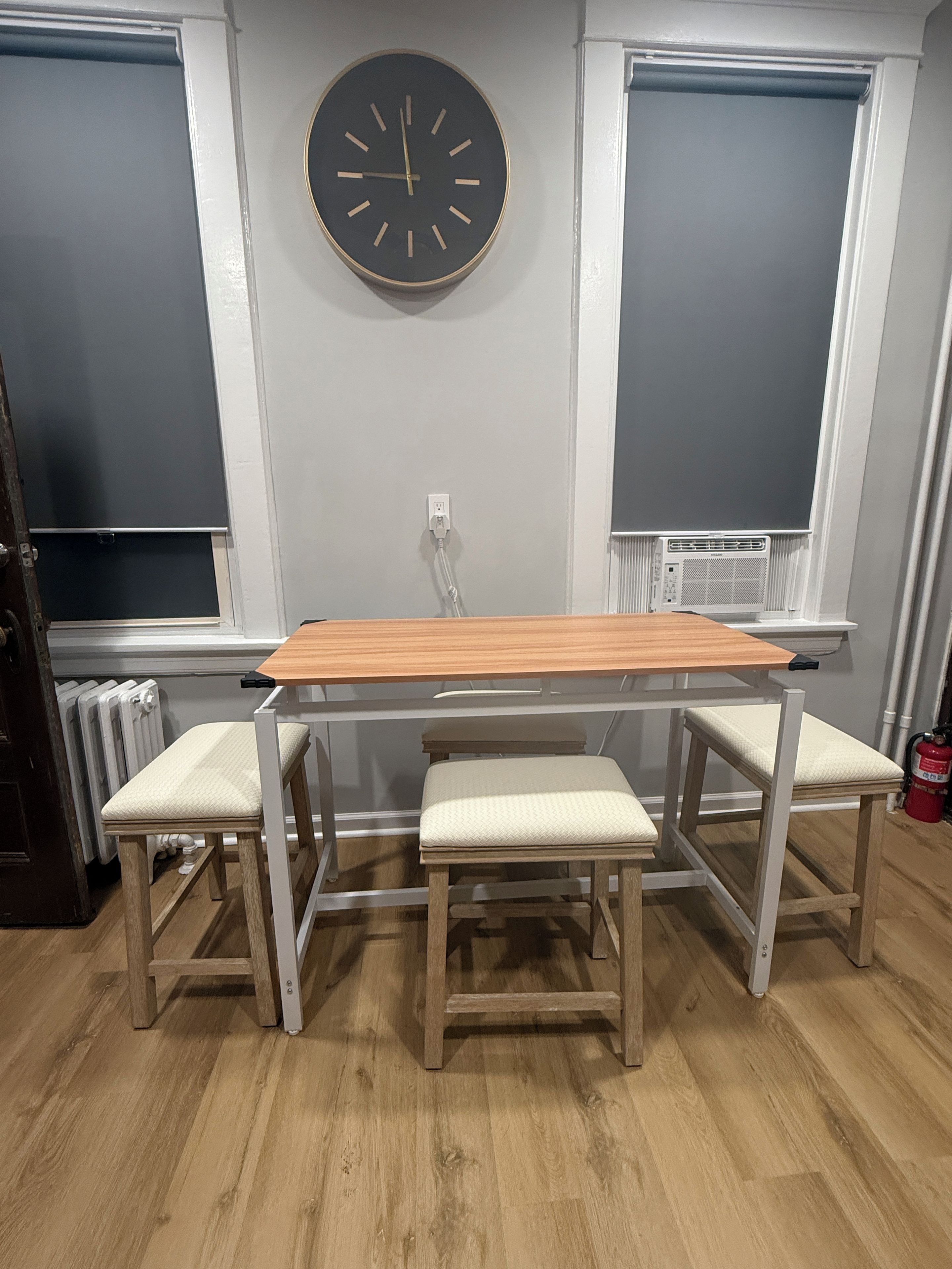 Ein Zimmer mit einem Holztisch, vier Stühlen, einer Wanduhr und zwei Fenstern mit Jalousien.