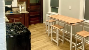 Uma cozinha com armários de madeira, fogão preto, mesa de jantar de madeira com quatro cadeiras, ventilador de teto e um relógio de parede.
