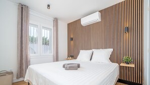 A modern bedroom with a large bed, wooden panel wall, air conditioning unit, bedside tables, and a view of the outdoors. - Pleasant house on the canal with pool (Castelló d'Empúries)