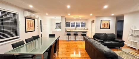 Una sala de estar moderna con una mesa de comedor de vidrio, un sofá de cuero negro y una zona de cocina al fondo.