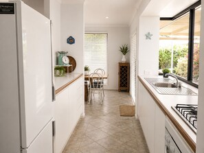 A modern kitchen with white cabinets, a stainless steel sink, and a gas stove. There is a dining area with a table and chairs, and a window with blinds.