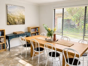 A dining area with a wooden table, white chairs, a bookshelf, and a window with blinds.
