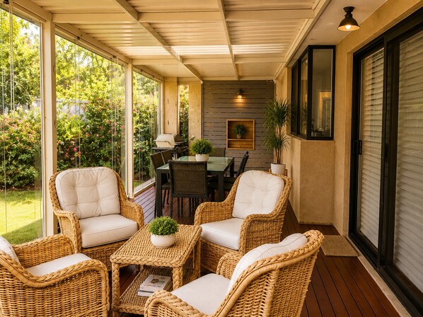 A covered patio with wicker furniture, a table, and chairs, a grill, and a view of a garden.