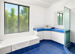 A bathroom with a bathtub, a sink with a blue countertop, and a large window with blinds.