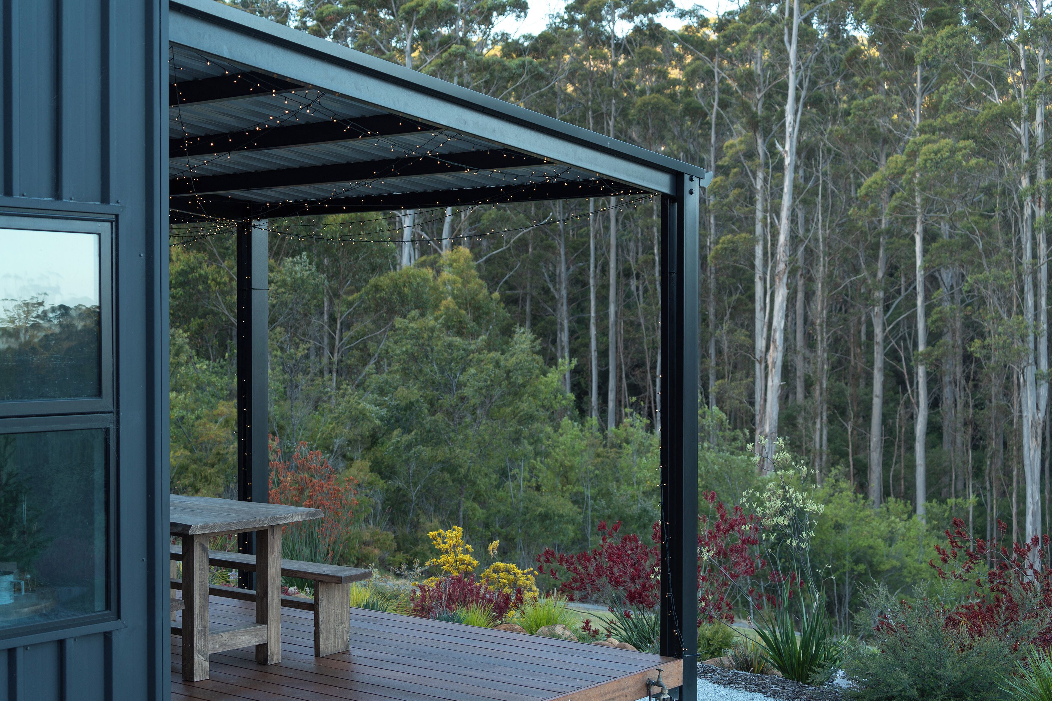 A wooden deck with a table and bench, surrounded by a garden with various plants and trees in the background.