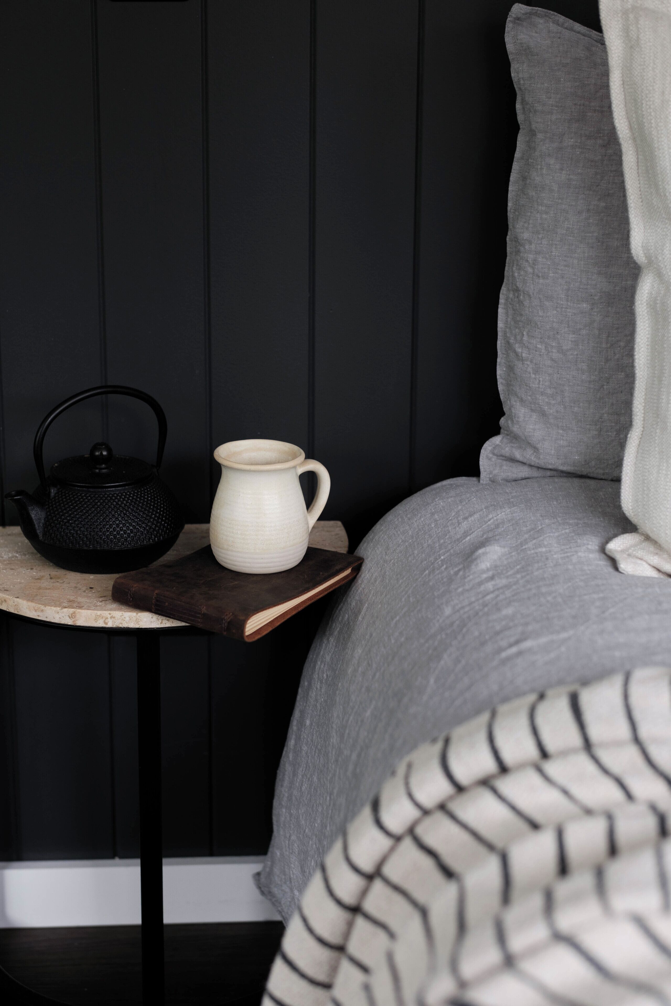 Un dormitorio ordenado con una cama, una mesita de noche con una tetera y una jofaina, y una pared oscura.