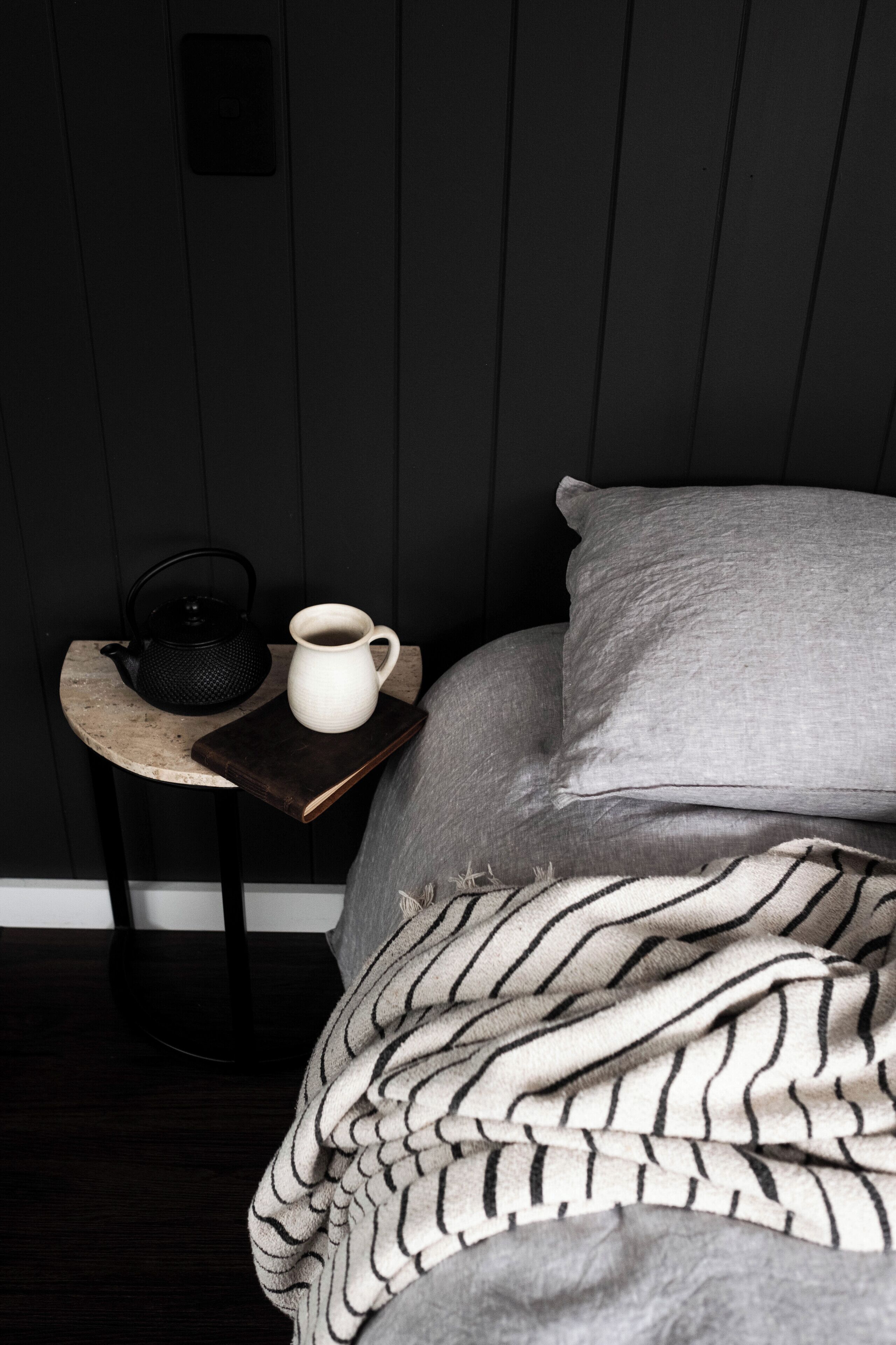 A bed with a grey blanket, a pillow, a white mug, and a black kettle on a wooden nightstand.