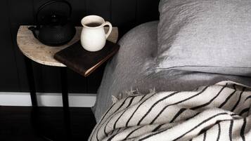 A bed with a grey blanket, a pillow, a white mug, and a black kettle on a wooden nightstand.