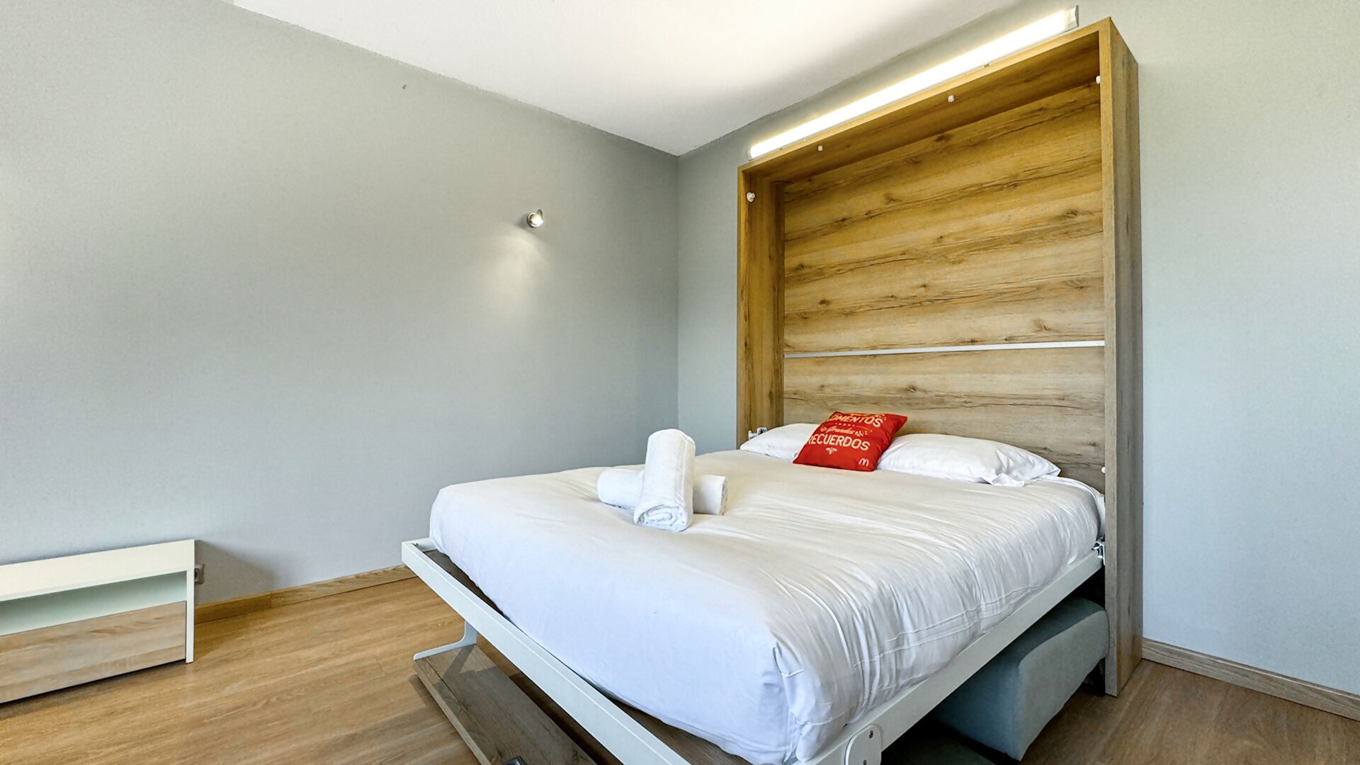 Une chambre d’hôtel moderne avec une tête de lit en bois, un lit bien fait avec du linge de lit blanc et un oreiller rouge.