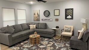 Ein modernes Wohnzimmer mit einem grauen Ecksofa, einem kleinen runden Holztisch, einem Teppich mit Blumenmuster und einem Deckenventilator.