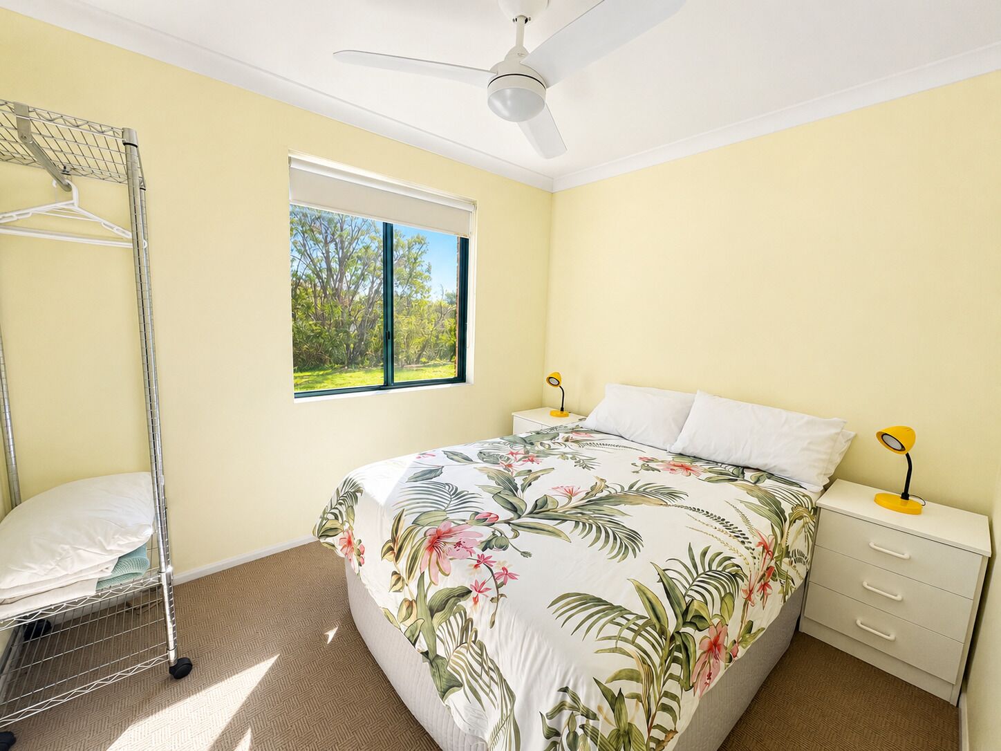 Quarto com cama, janela, ventoinha no teto, mesa de cabeceira, abajur e um suporte para roupas.