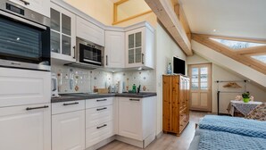 Een moderne keuken met witte kasten, een magnetron en een inbouwoven. Aan de rechterkant een houten kast en op de voorgrond een bed met een blauw beddegoed met patroon.