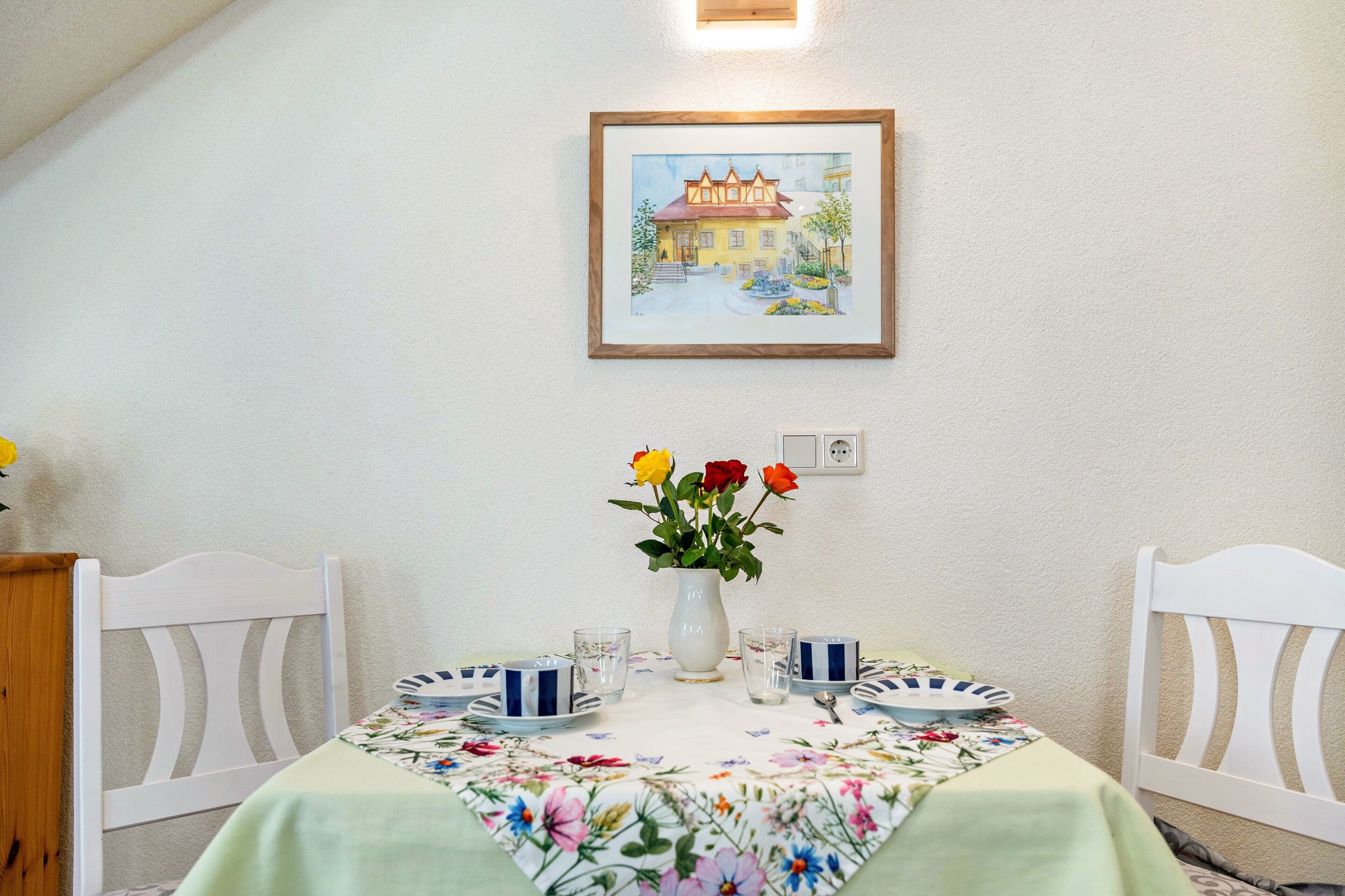 Een eethoek met een tafel gedekt voor twee personen, een lijst met een schilderij erboven en een vaas met bloemen aan de muur.