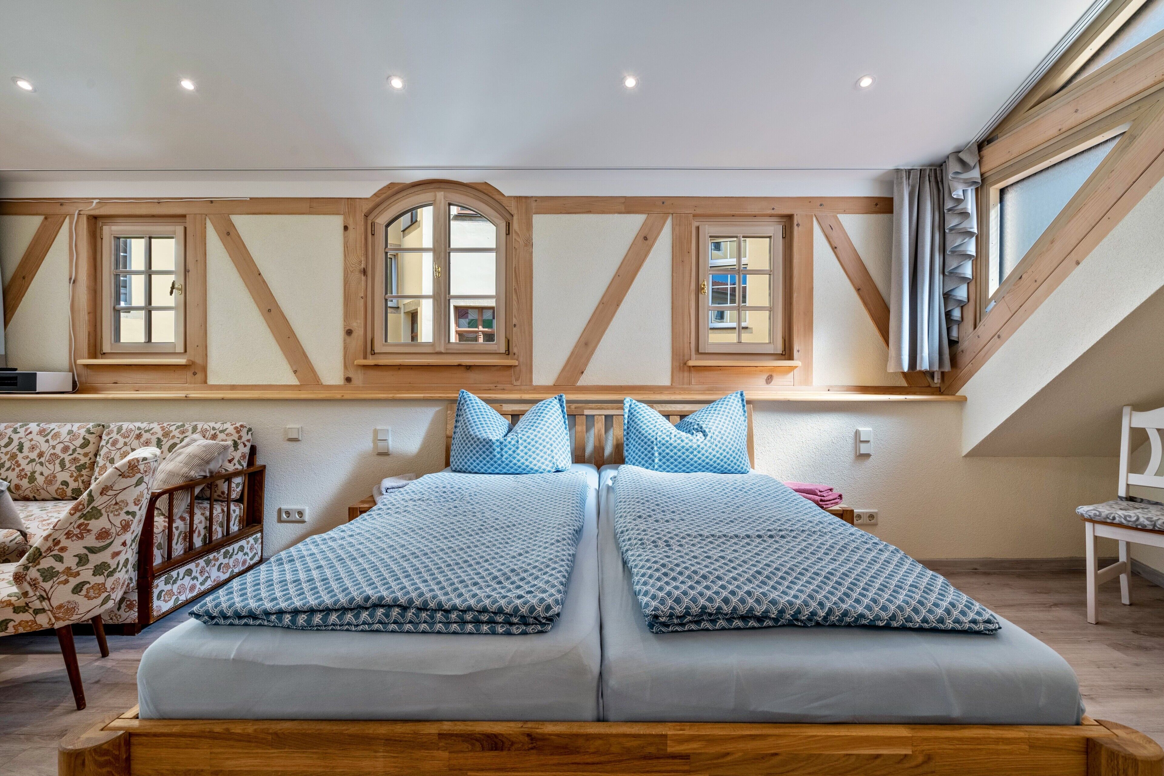A bedroom with a wooden headboard, two beds with patterned bedding, a floral armchair, and a window with a view.