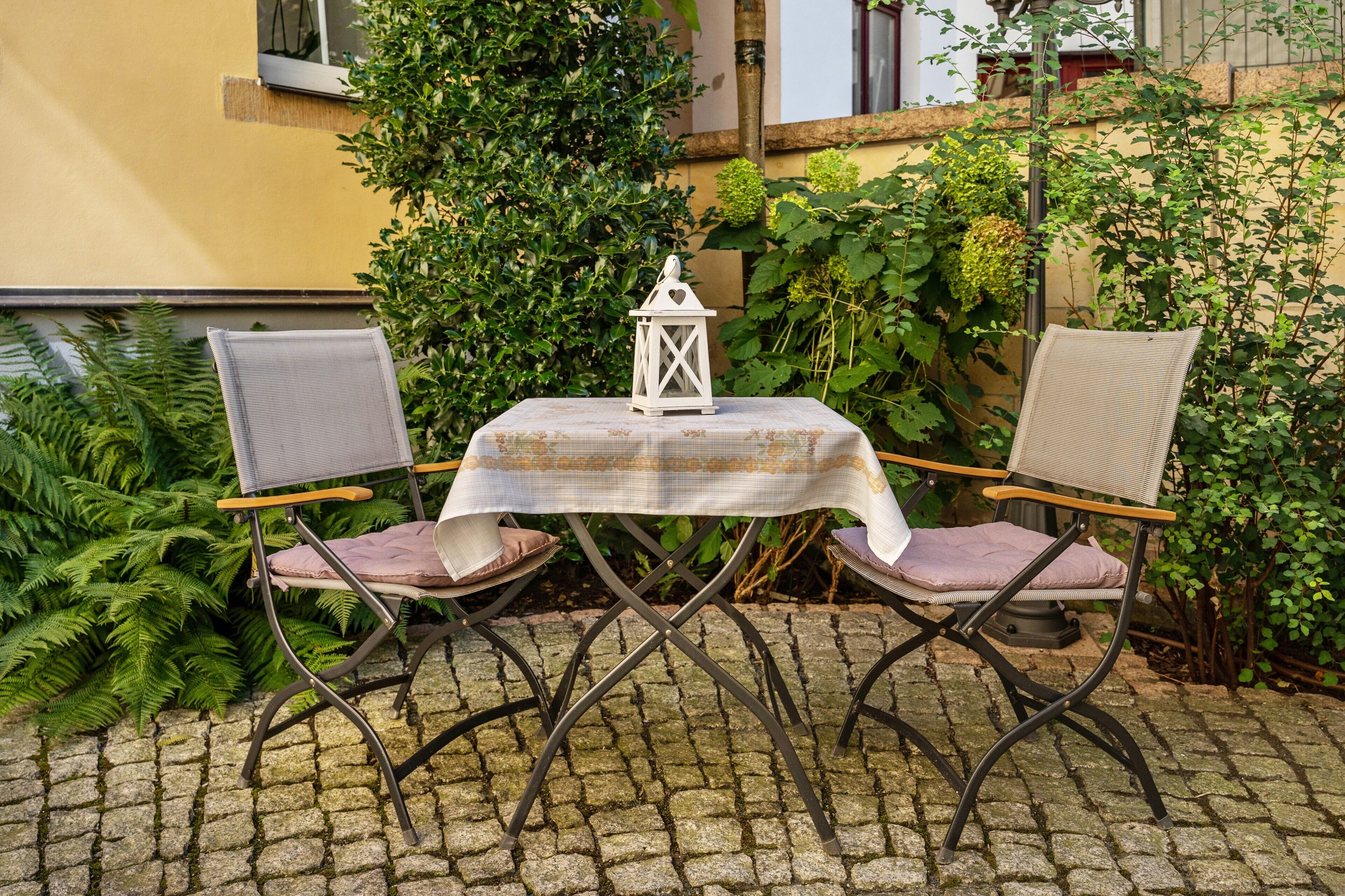Un espace extérieur aménagé avec deux chaises et une table, entouré de verdure et d’un bâtiment.
