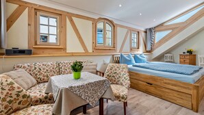 Een slaapkamer met een schuine plafond, houten balken, een bed met blauw beddengoed, een tafel met een groene plant, en zitplaatsen met een bloemenprint.