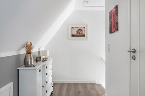 Un couloir avec une commode blanche, un tableau encadré au mur et une porte.