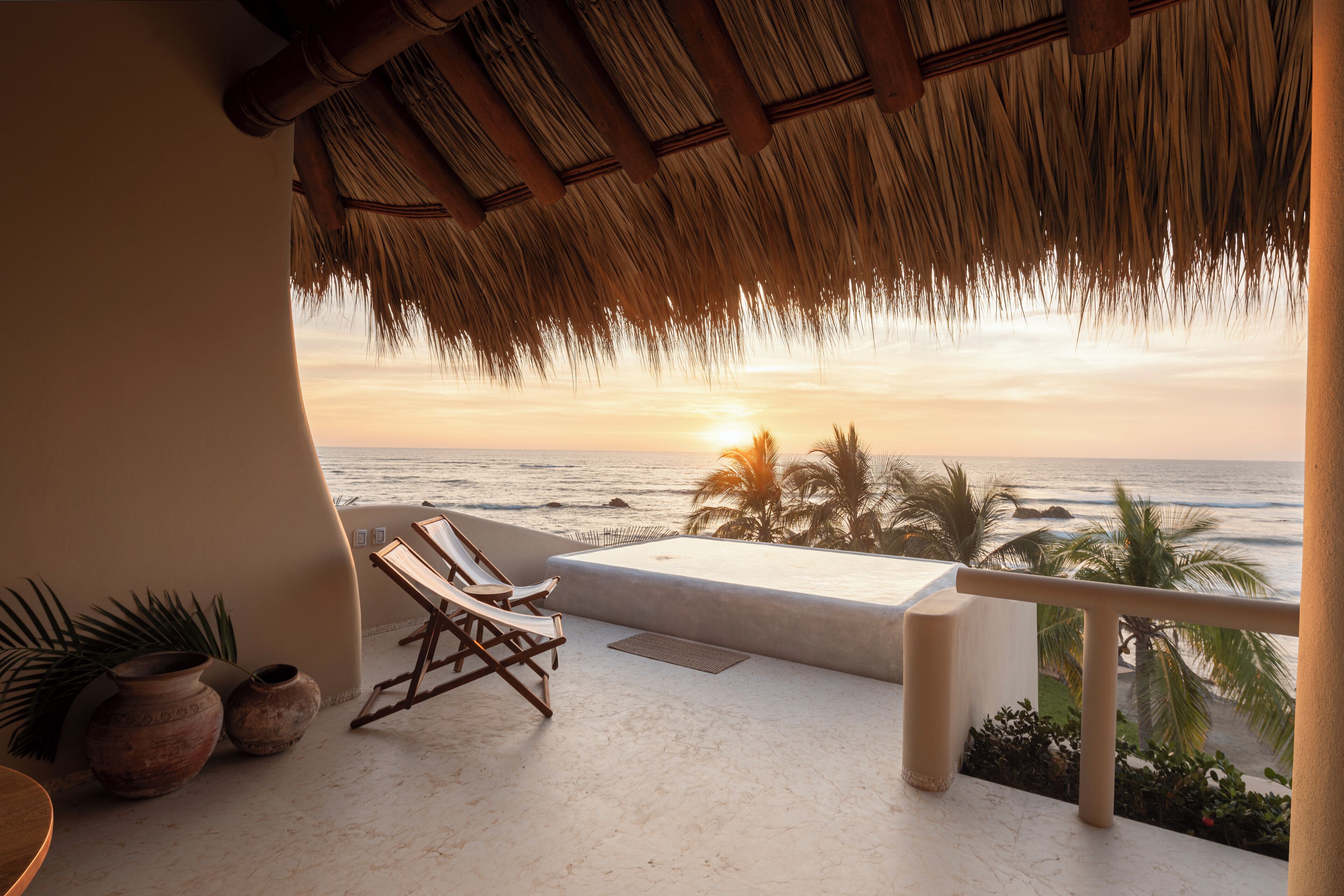 Un balcon offrant une vue sur l’océan au coucher du soleil, un toit de chaume et un espace salon avec des chaises.