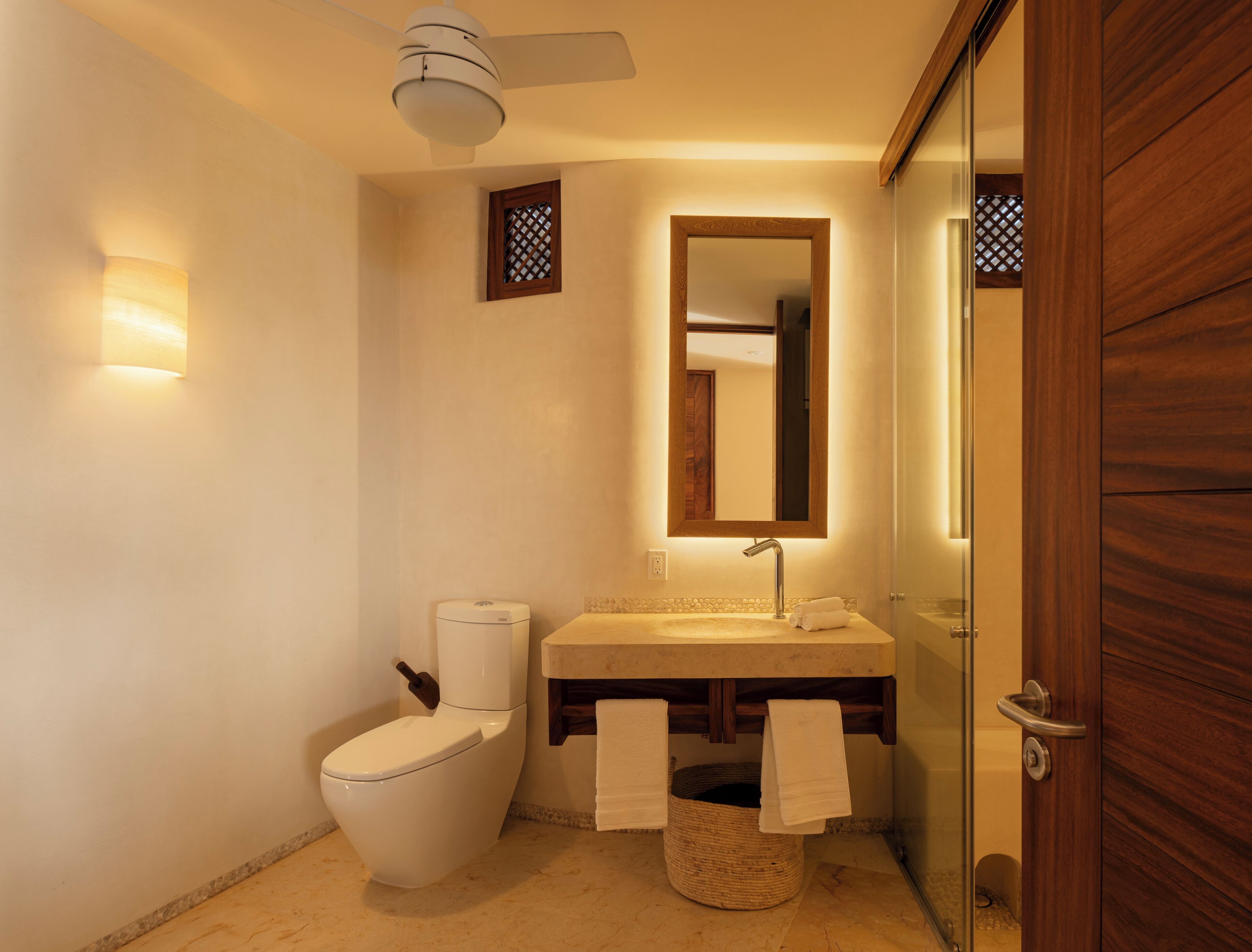 Ein modernes Badezimmer mit Toilette, Waschbecken und Spiegel. Ein Deckenventilator und ein kleines Fenster mit Gittermuster sind vorhanden.