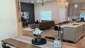 Une cuisine moderne dotée d’un îlot central, un coin repas avec une longue table et des chaises, et un espace de vie avec un canapé et un téléviseur.