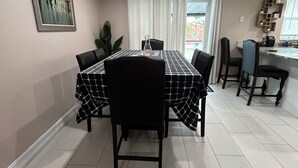 Uma área de refeições com mesa posta para quatro pessoas, rodeada por cadeiras, em um cômodo com piso ladrilhado e iluminação embutida no teto.