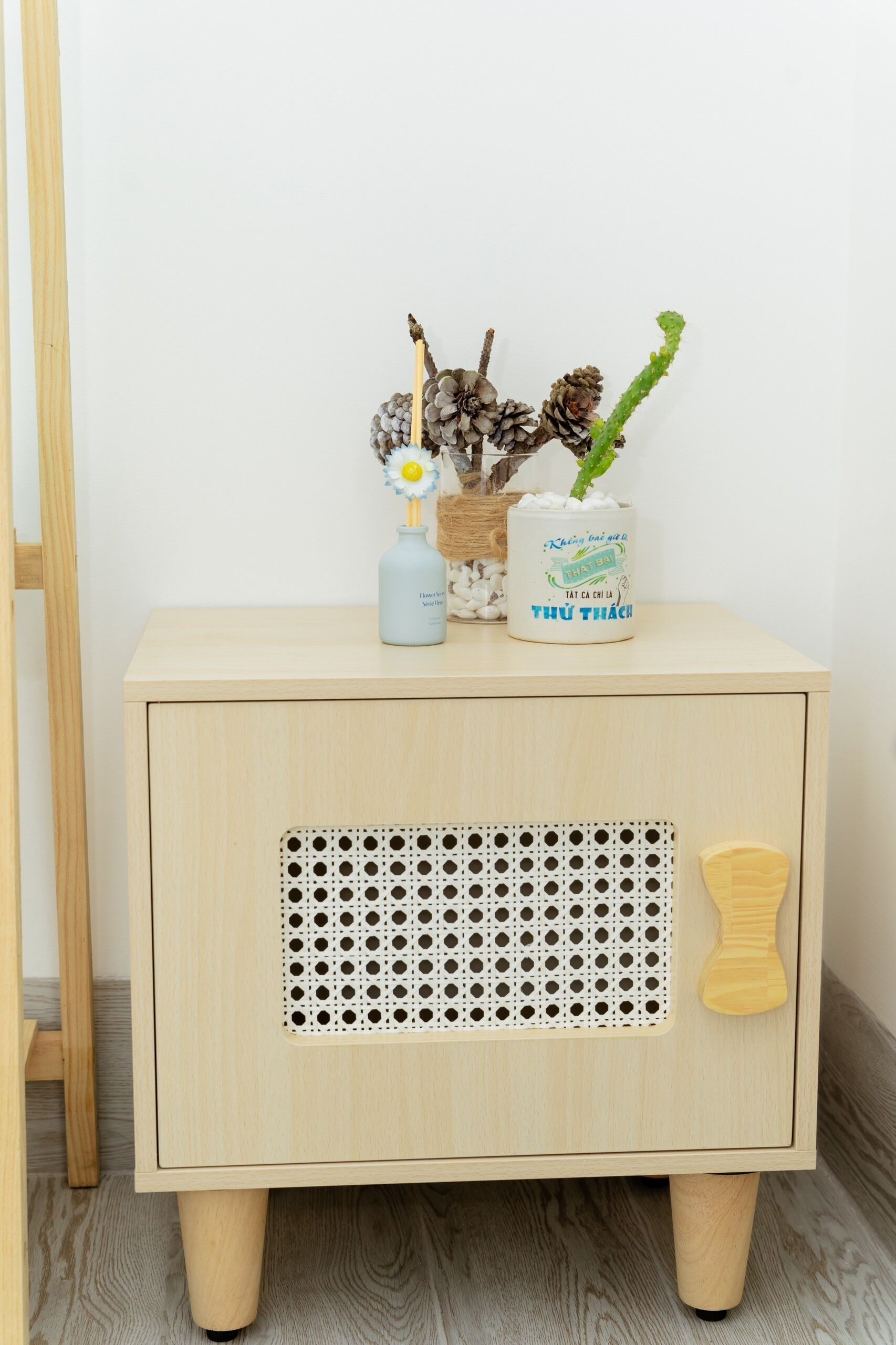 A wooden nightstand with a perforated panel and a drawer, topped with decorative items including a vase, a bottle, and pinecones.