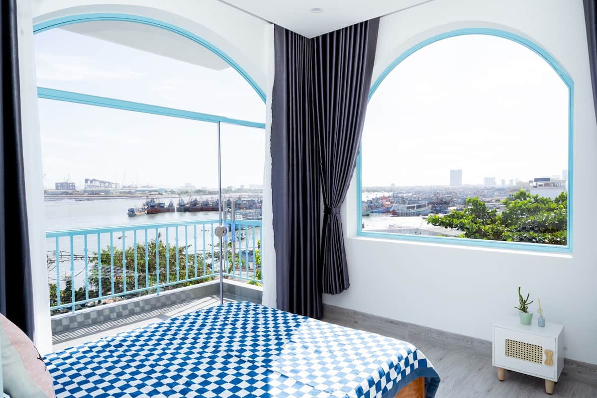 Une chambre avec une grande fenêtre, un lit recouvert d’une couverture à carreaux, une table de chevet, et une vue sur une rivière et le paysage urbain.