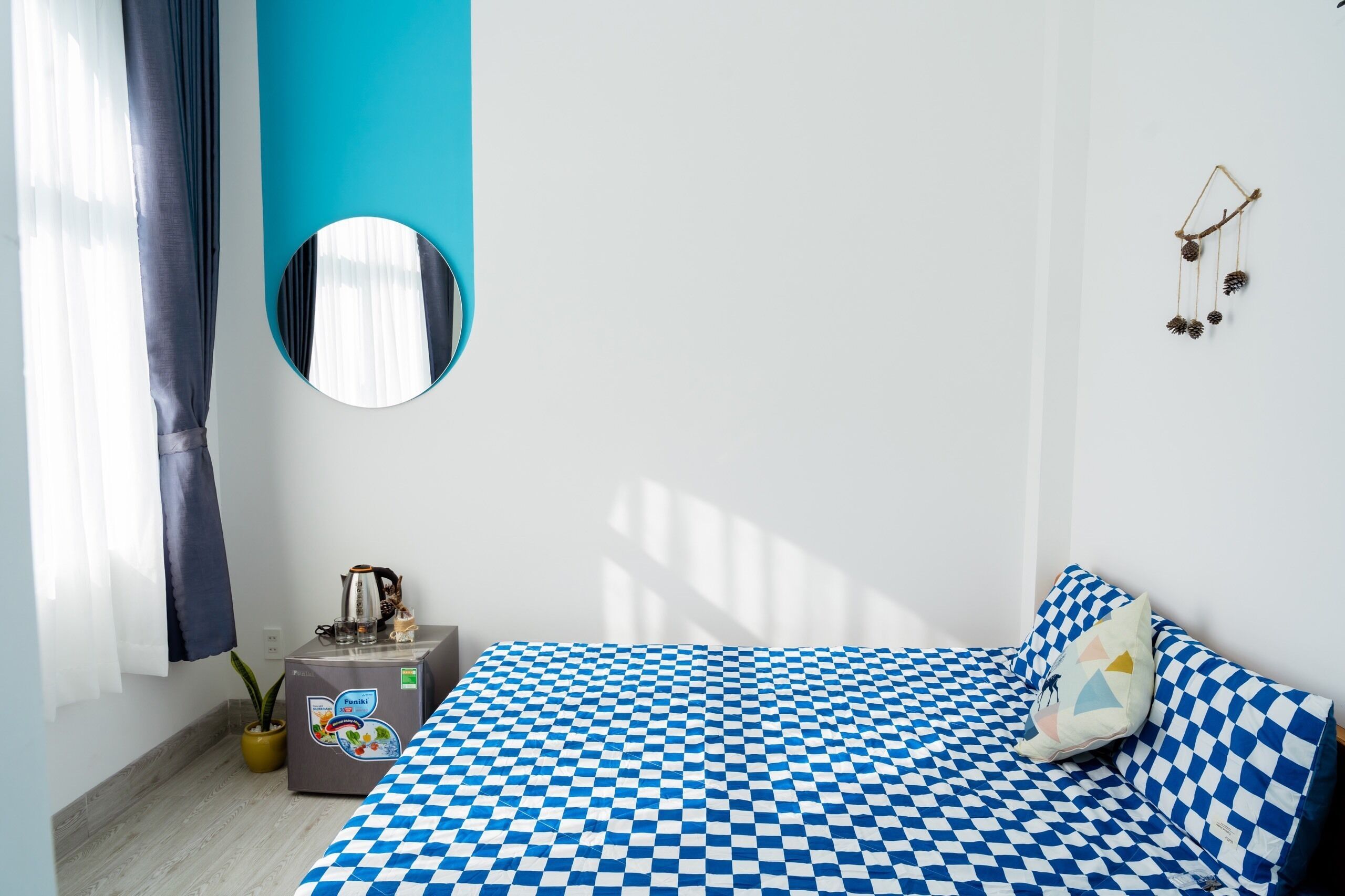 Een slaapkamer met een blauw-wit geruite spreidekbed, een klein nachtkastje met een koffiezetapparaat, een potplant en een spiegel aan de muur.