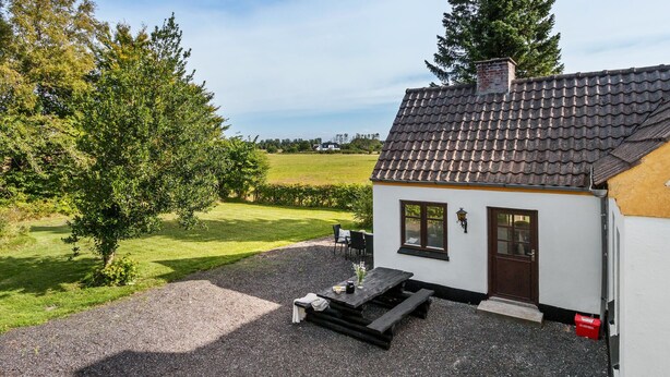 A small white house with a tiled roof, a wooden bench, and a table with a vase and a cup. - Lovely home in Dronninglund with WiFi (Dronninglund)