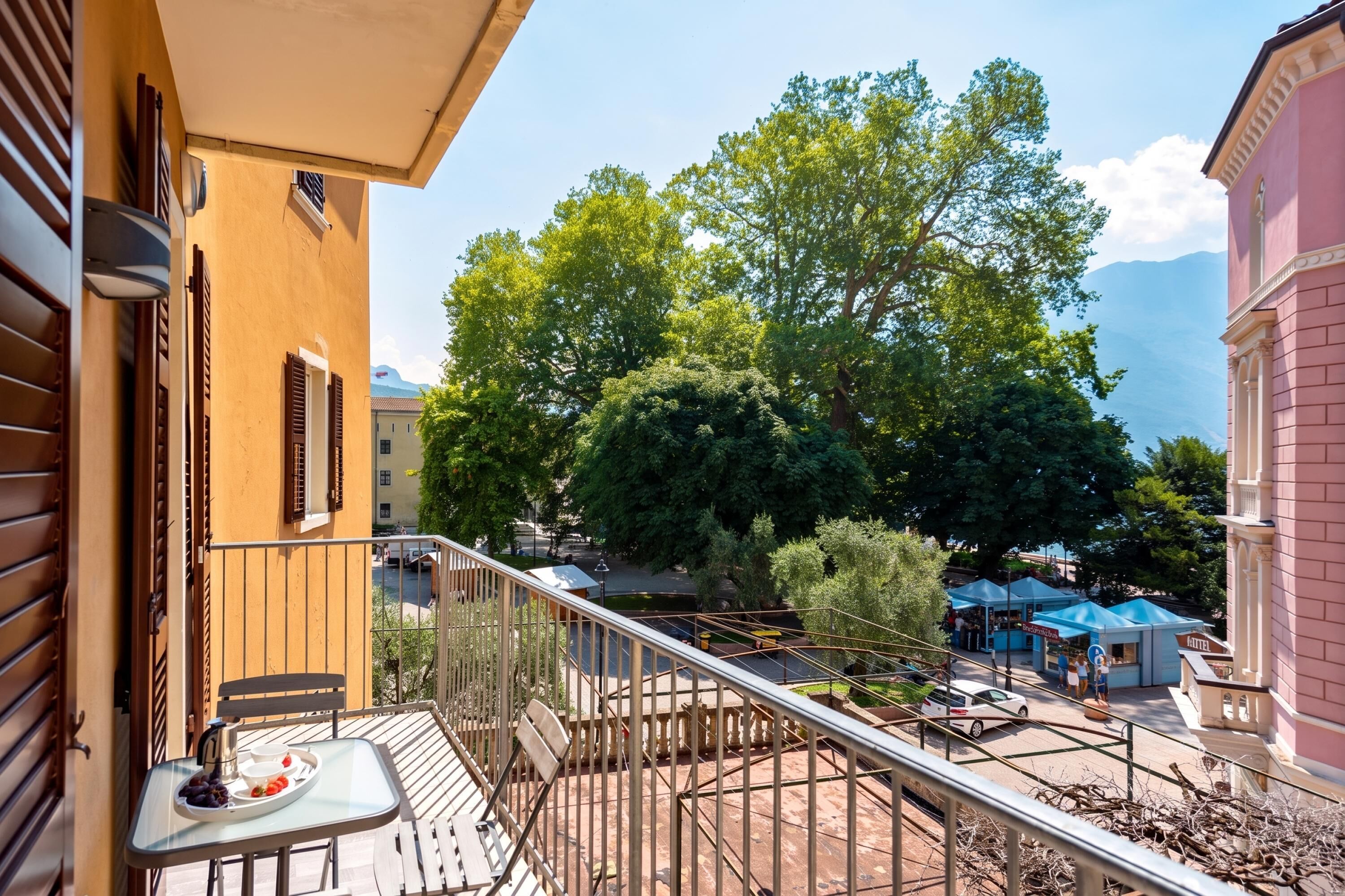 A balcony with a table set for two, overlooking a courtyard with trees and a view of a mountain range.