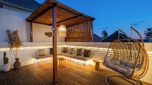 A wooden deck with a cushioned seating area, a hanging chair, and a wall-mounted lamp. - Villa Dove By OriVista (Canggu)