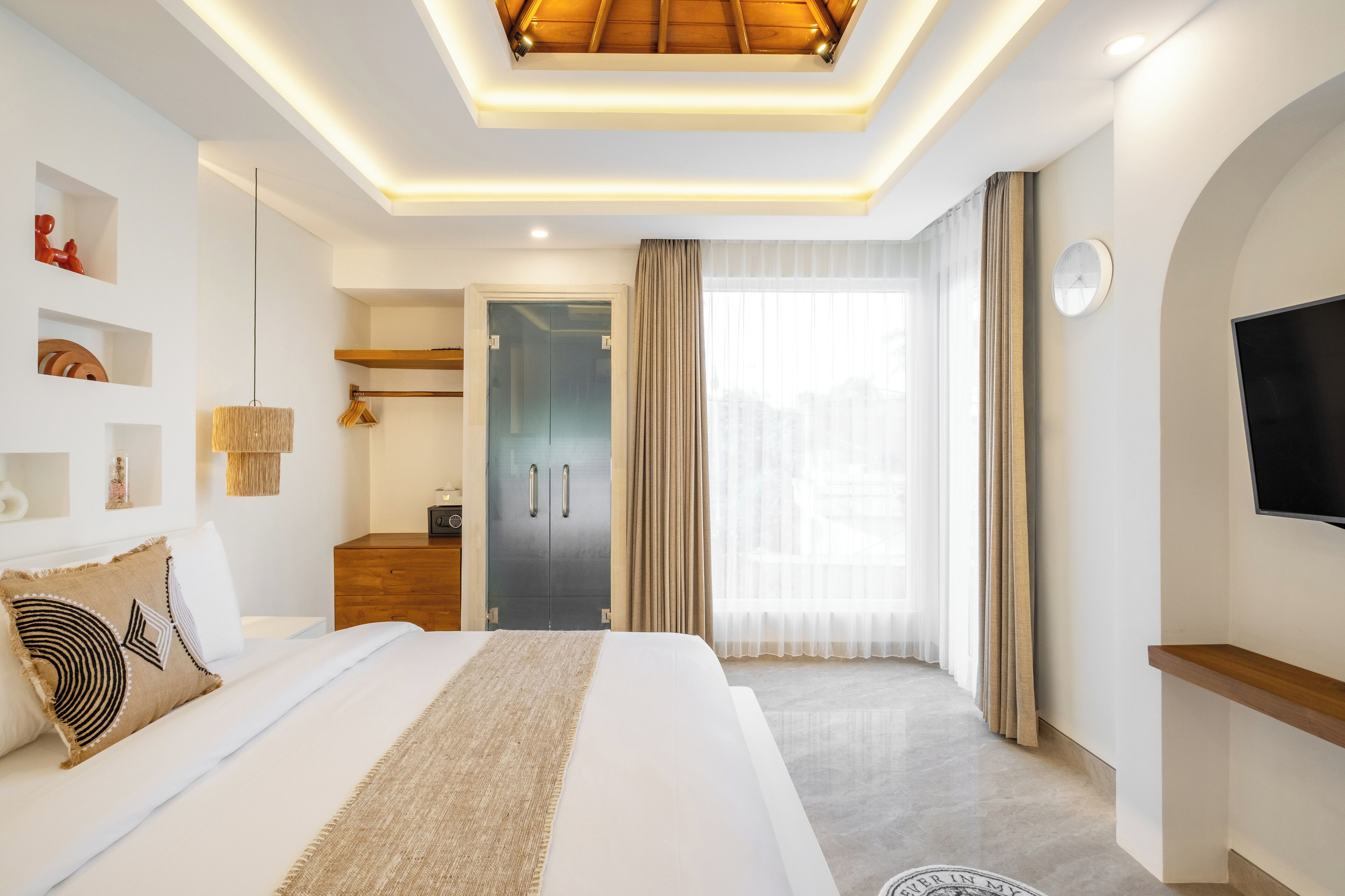Ein modernes Hotelzimmer mit einem großen Bett, einem Fernseher, einem Kleiderschrank und einem durch eine Glastür einsehbaren Badezimmer.