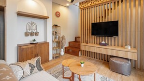 A modern living room with a wooden ceiling, a flat-screen TV mounted on a wooden panel wall, a wooden cabinet, a small staircase, and a round wooden coffee table with a plant. - Villa Dove By OriVista (Canggu)