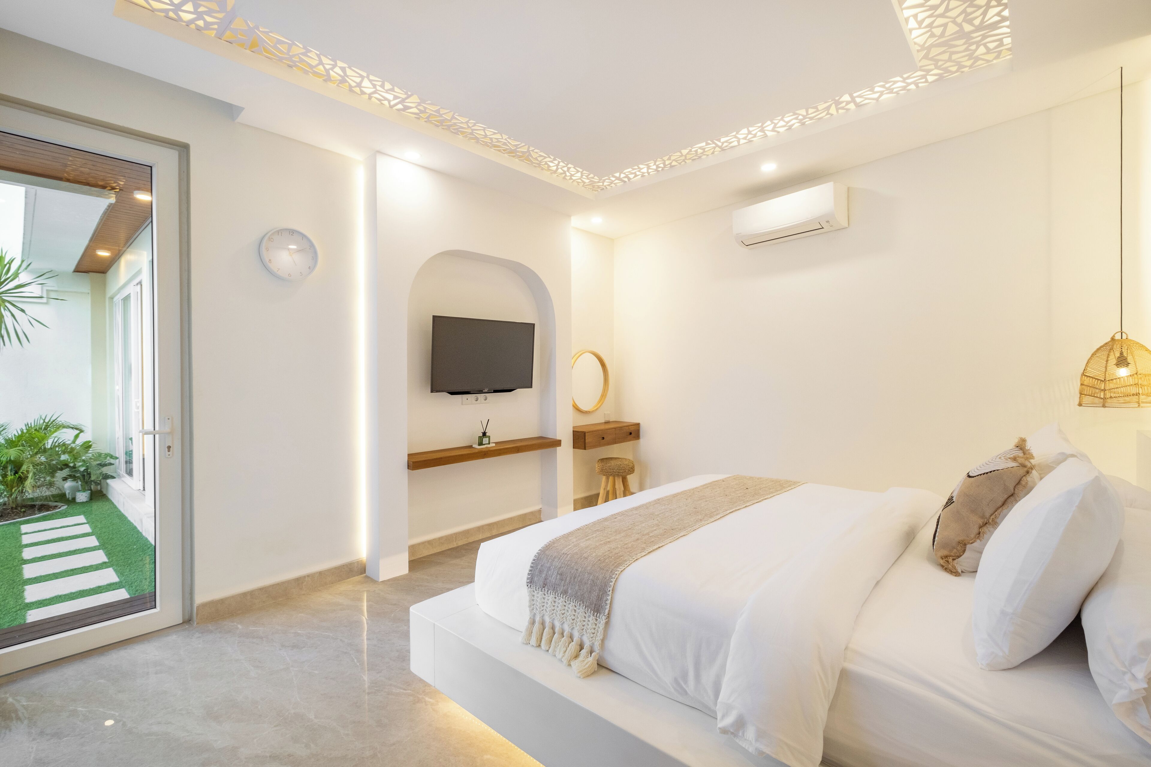 A modern bedroom with a large bed, a TV mounted on the wall, a small seating area, and a view of a green outdoor area.