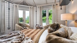 Ein Zimmer mit einem großen fellbezogenen Bett, einem Nachttisch mit Gläsern und einer Zeitschrift und Blick auf Grünflächen.