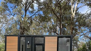 Una cabaña moderna y compacta con fachada de madera, ventanas amplias y una pequeña terraza con sillas.