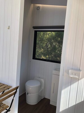 A compact bathroom with a toilet, a small cabinet, and a window with a view of trees. - Sandy Strait Shack by Tiny Away (Booral)