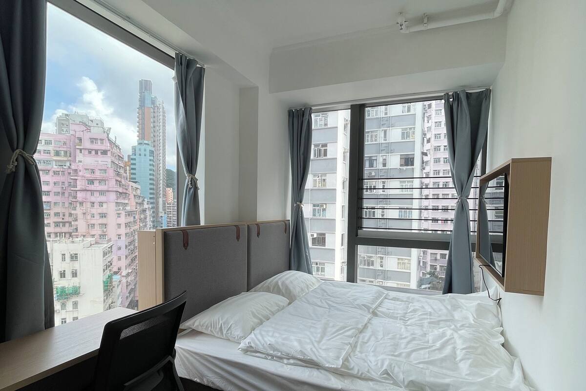 A bedroom with a bed, a desk, and a chair. There is a window with curtains and a view of buildings outside.