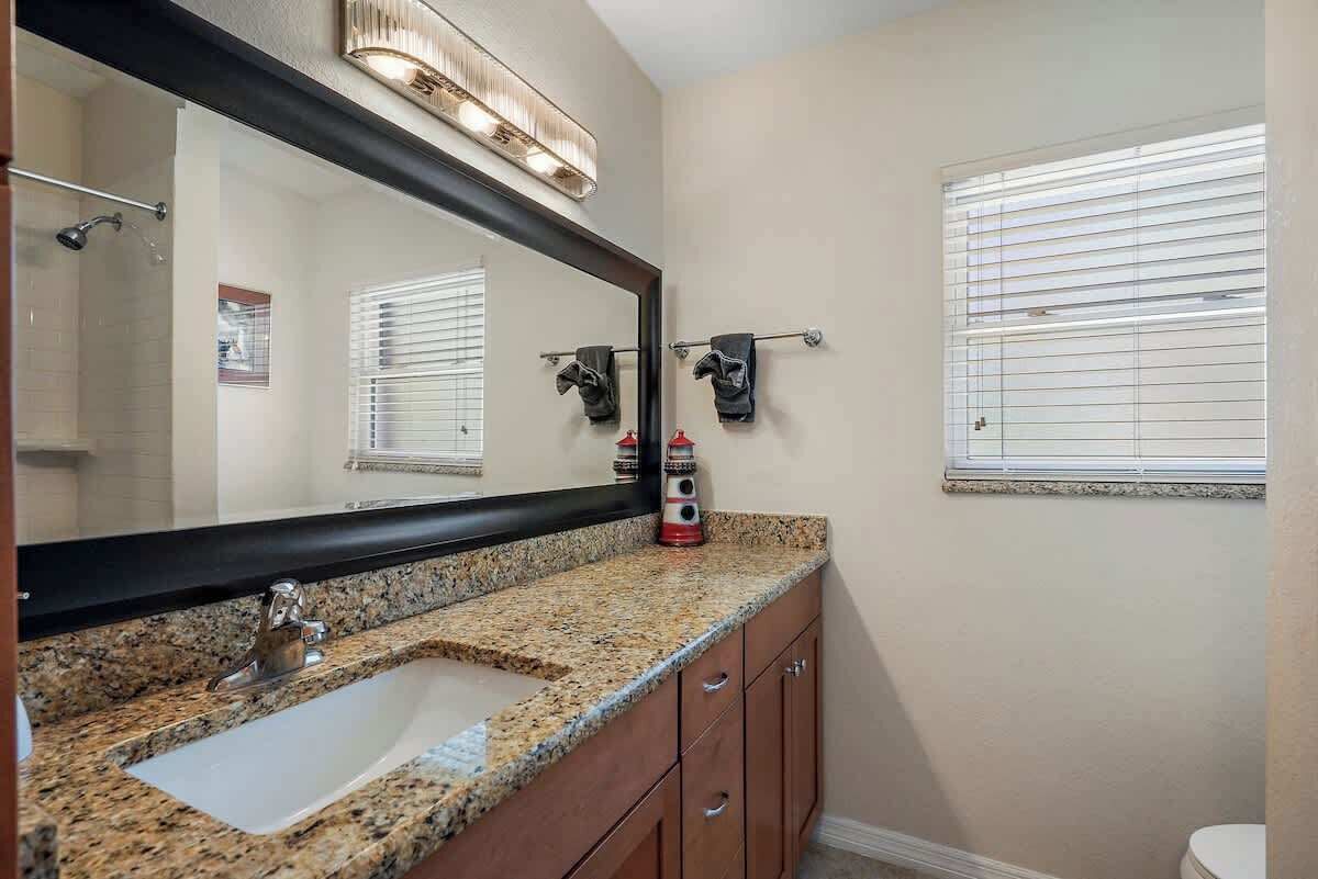 A bathroom with a granite countertop sink, a large mirror, a towel rack, and a window with blinds.