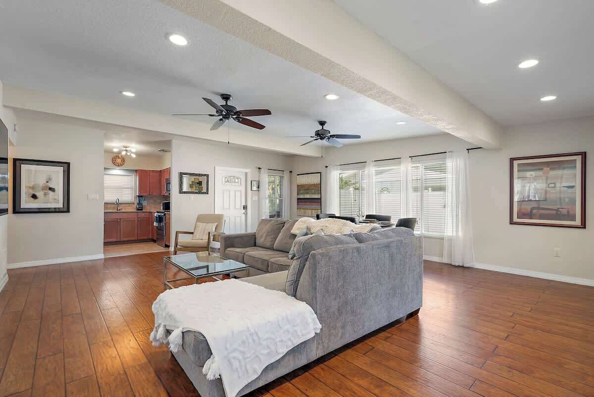 A spacious living room with a gray sofa, a coffee table, and a white throw blanket. There are two ceiling fans and a kitchen in the background.