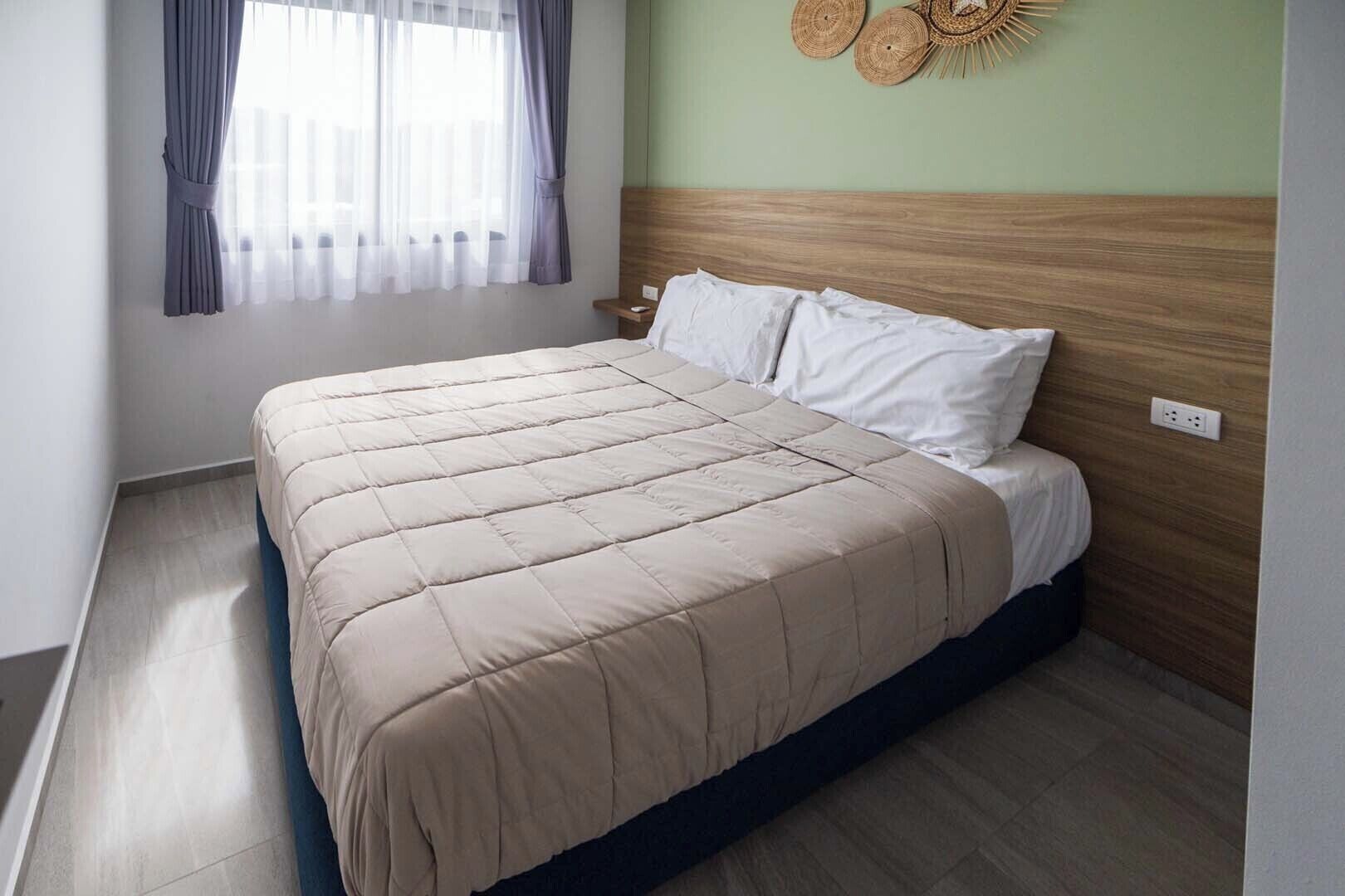 A neatly made bed with a beige quilt, a wooden headboard, and a wall-mounted light fixture.