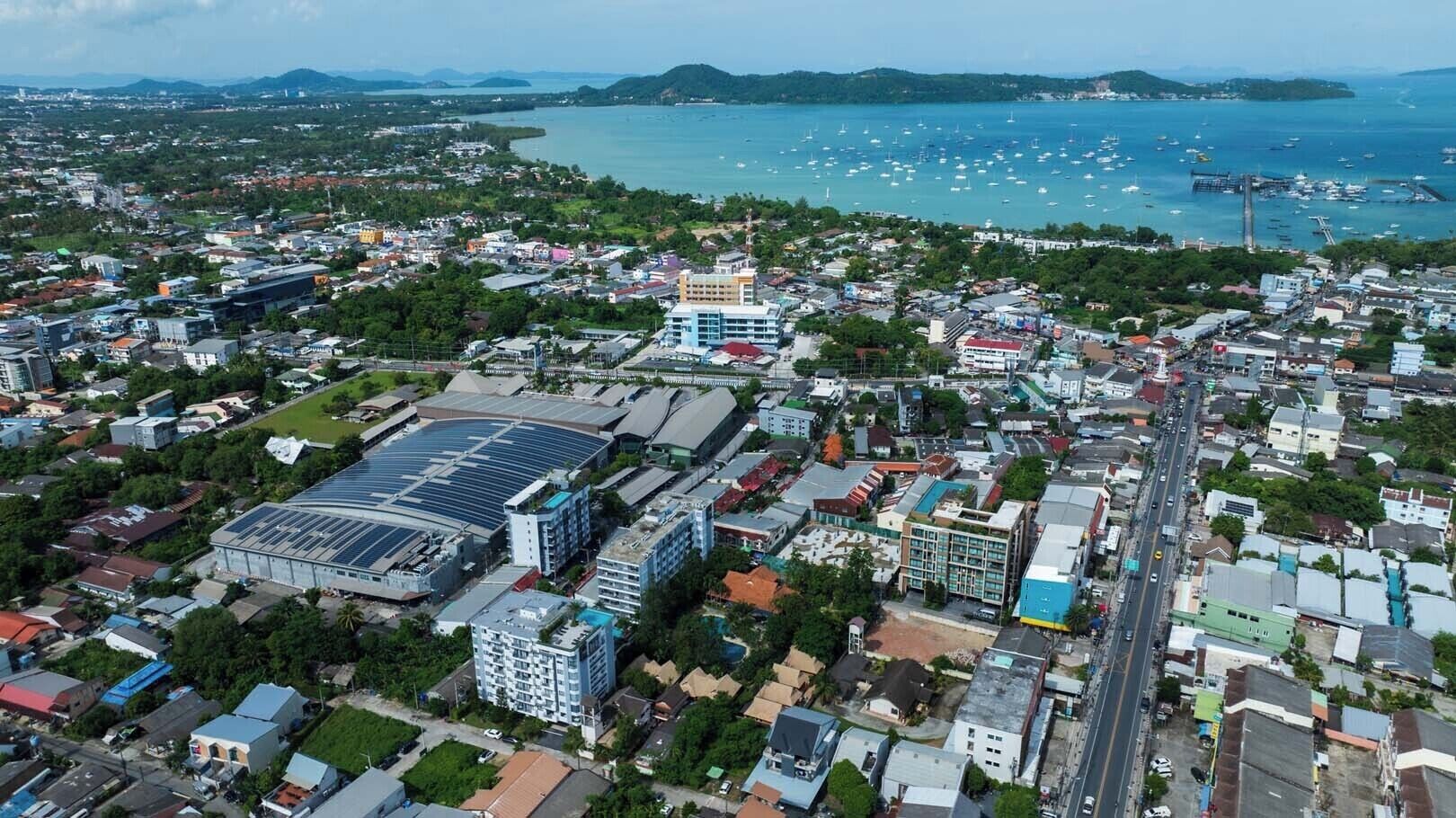 Vista aérea de una ciudad costera con una combinación de edificios residenciales y comerciales, una amplia instalación de energía solar y un puerto deportivo con numerosos barcos.