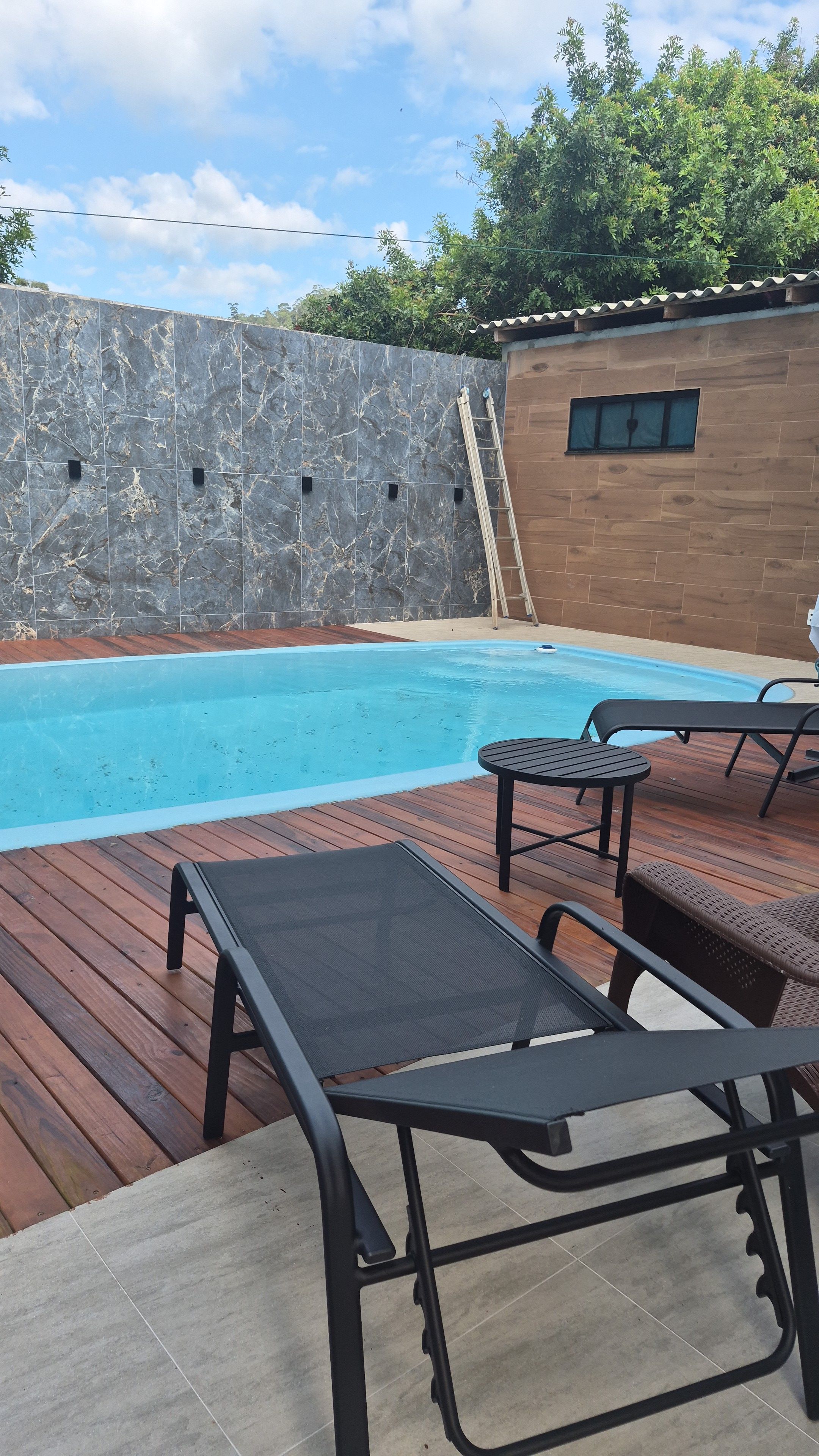 Ein Schwimmbecken mit Holzterrasse, Liegestühlen und einer Holzverkleidung mit einem Fenster.