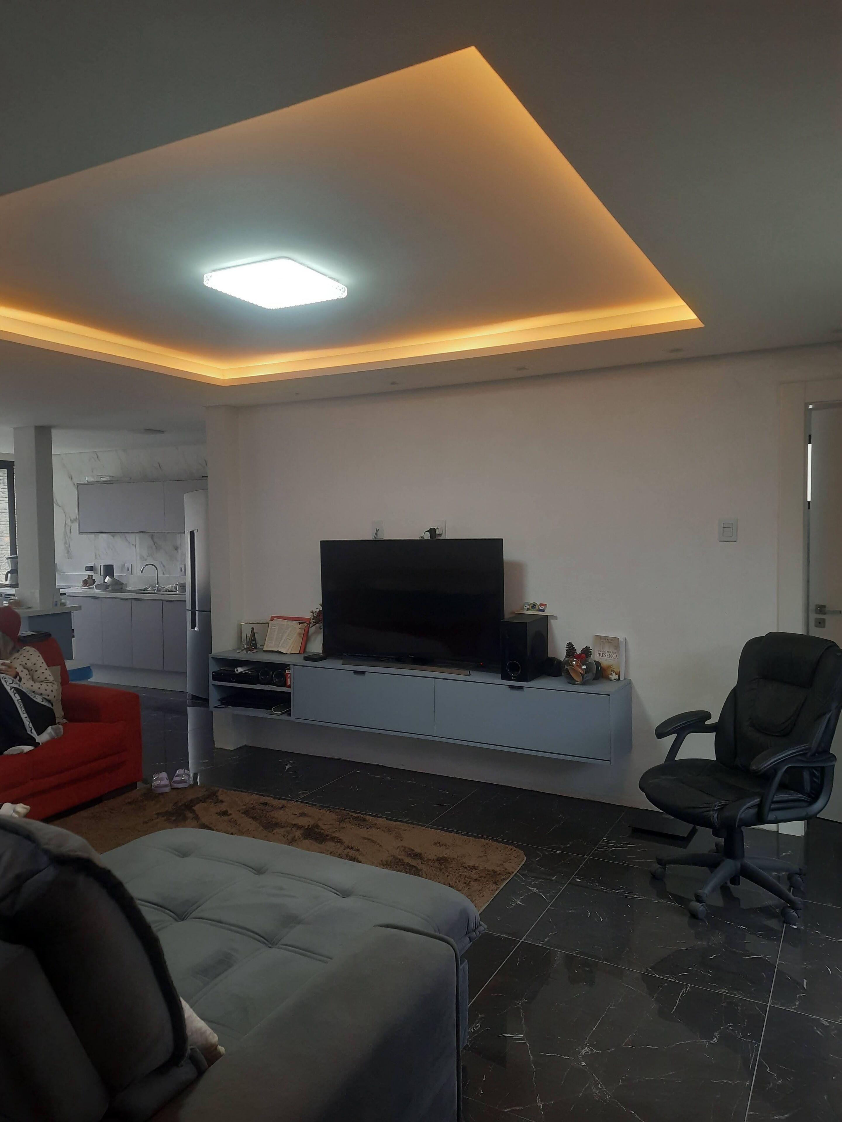 Een moderne woonkamer met een flatscreen televisie op een wandplank, een rode bank, een grijze zithoek en een zwarte bureaustoel.