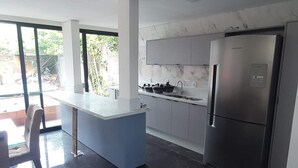 A modern kitchen with a built-in refrigerator, a dining area with a blue island, and a large window providing a view of the outdoors. - Beach House Delegado Marcio (Praia do Estaleiro Balneário Camboriú)